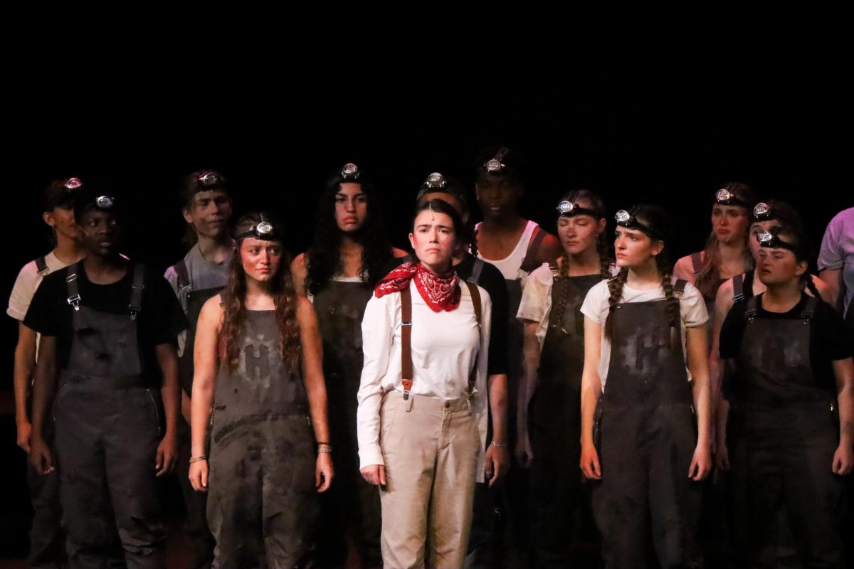 Lila Gibbs, playing Orpheus, stands on stage, surrounded by members of the ensemble. 