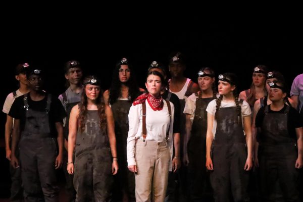Lila Gibbs, playing Orpheus, stands on stage, surrounded by members of the ensemble. 